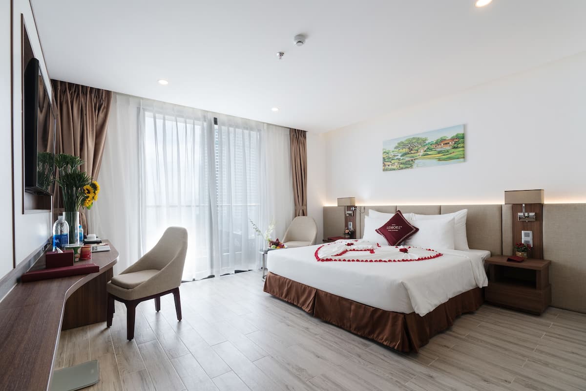 A hotel room with a large bed, a desk, and a chair. There is a painting on the wall and a window with curtains.
