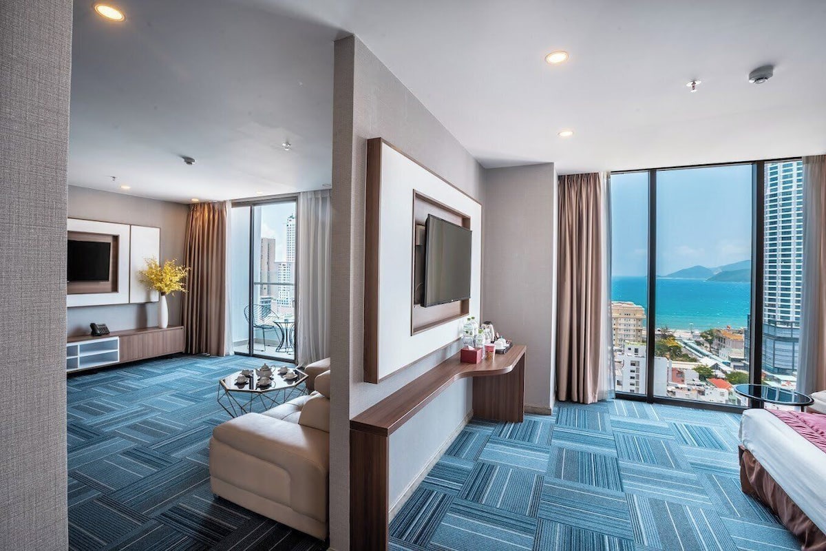 A modern hotel room with a large bed, a TV, a seating area with a table and chairs, a small desk, and a balcony with a city view.