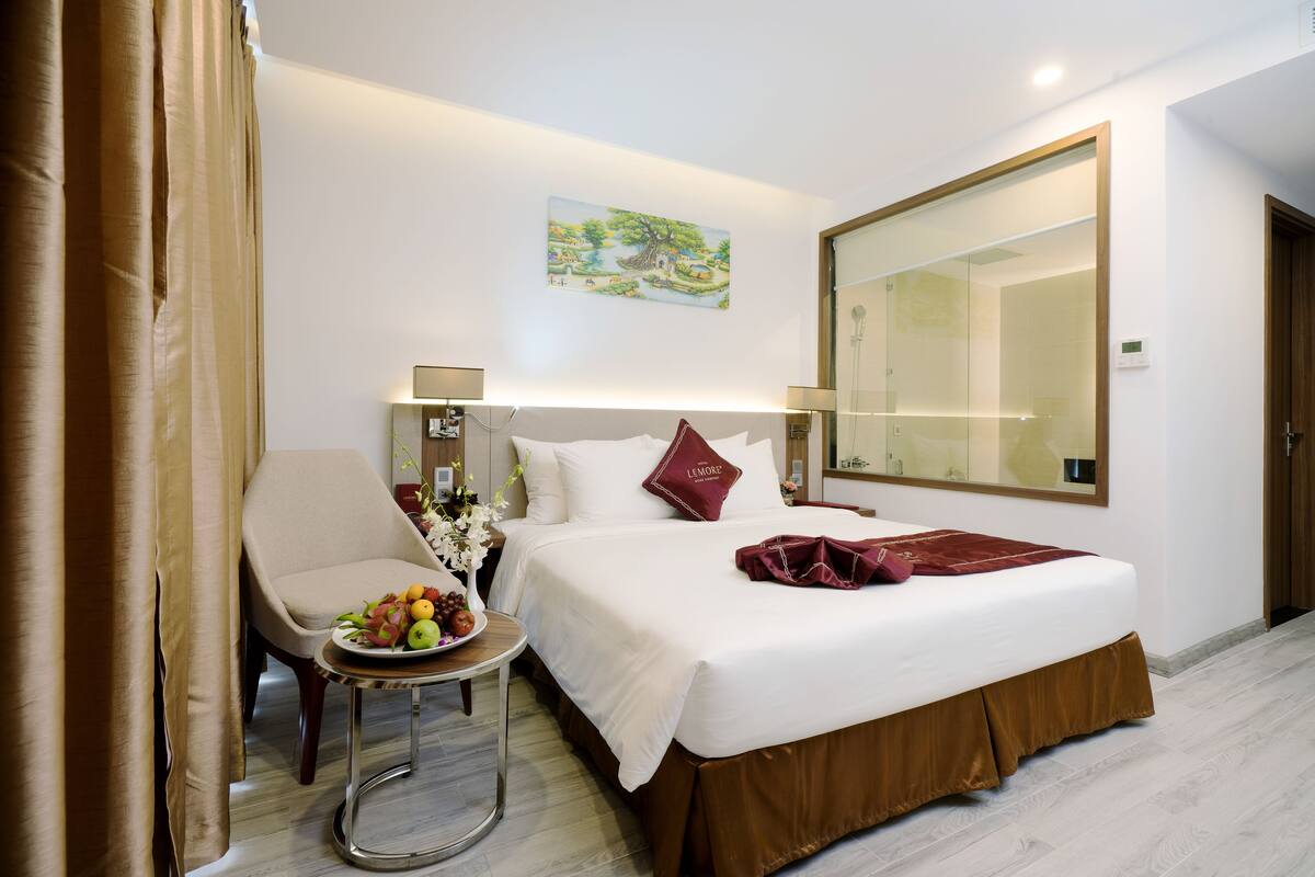 A hotel room with a bed, a chair, a small table with fruit, and a painting on the wall.