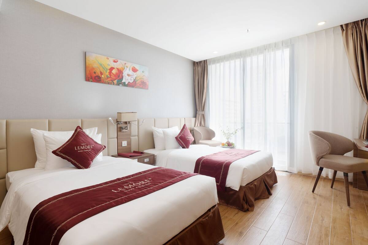 A hotel room with two beds, a desk, a chair, and a large window with curtains.
