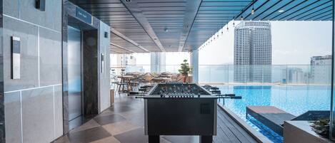 A modern indoor pool area with a foosball table, a glass-enclosed pool, and a view of high-rise buildings.