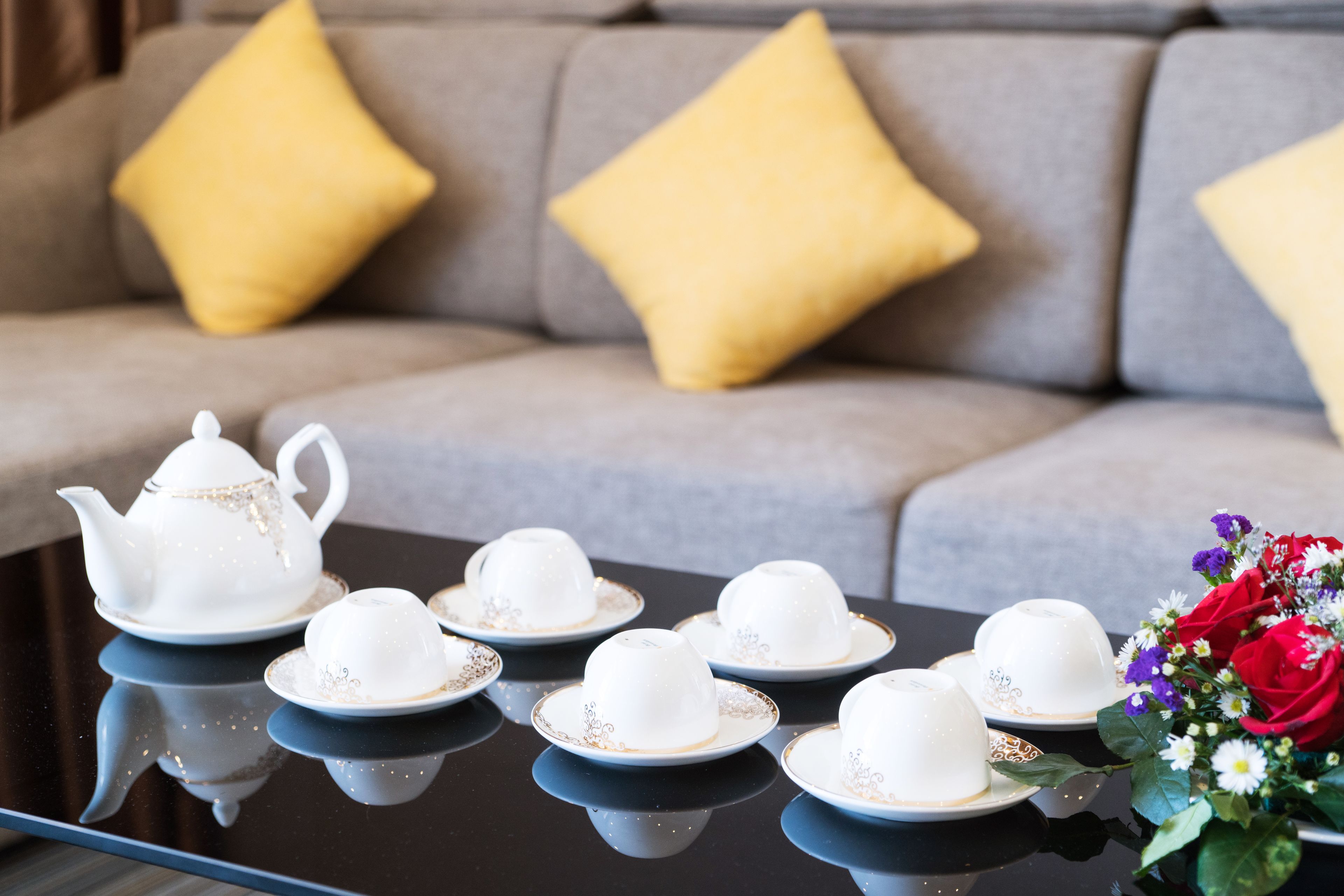 A table with white teapots and cups, a sofa with yellow cushions, and a bouquet of flowers.