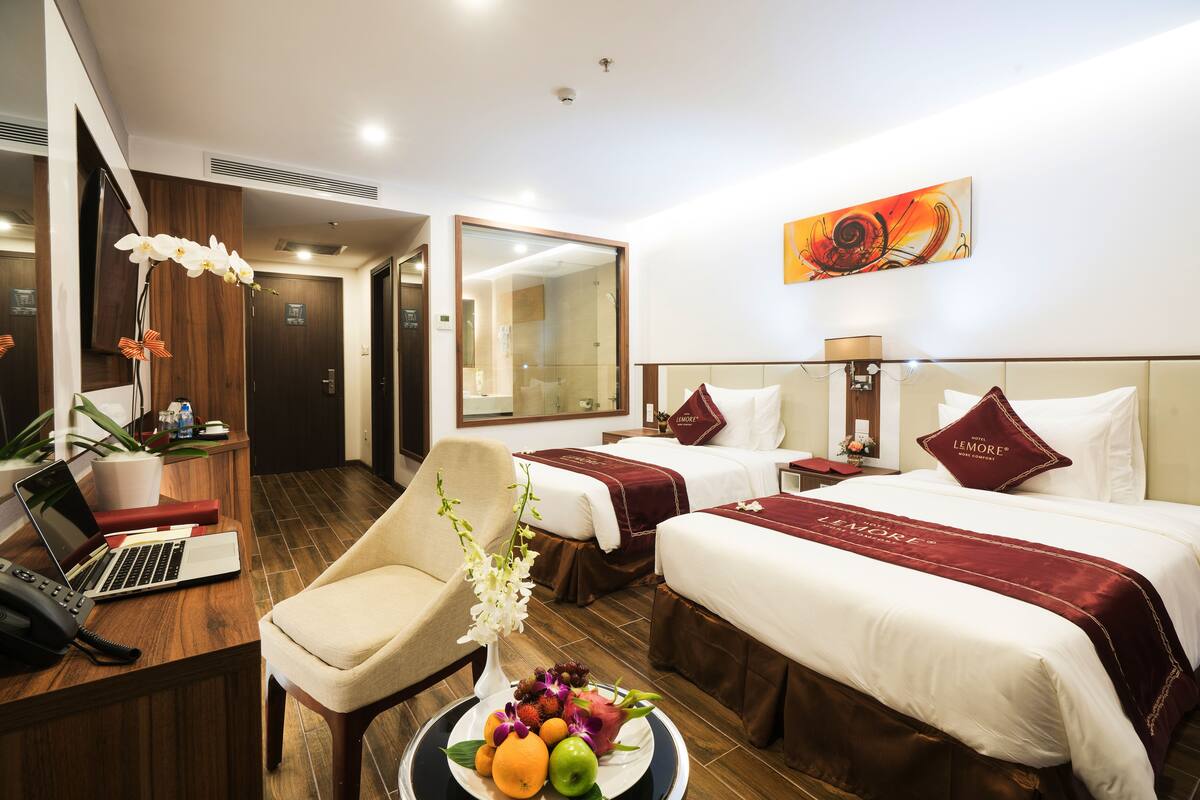 A hotel room with two beds, a desk with a laptop, a chair, a fruit bowl, and a large mirror.