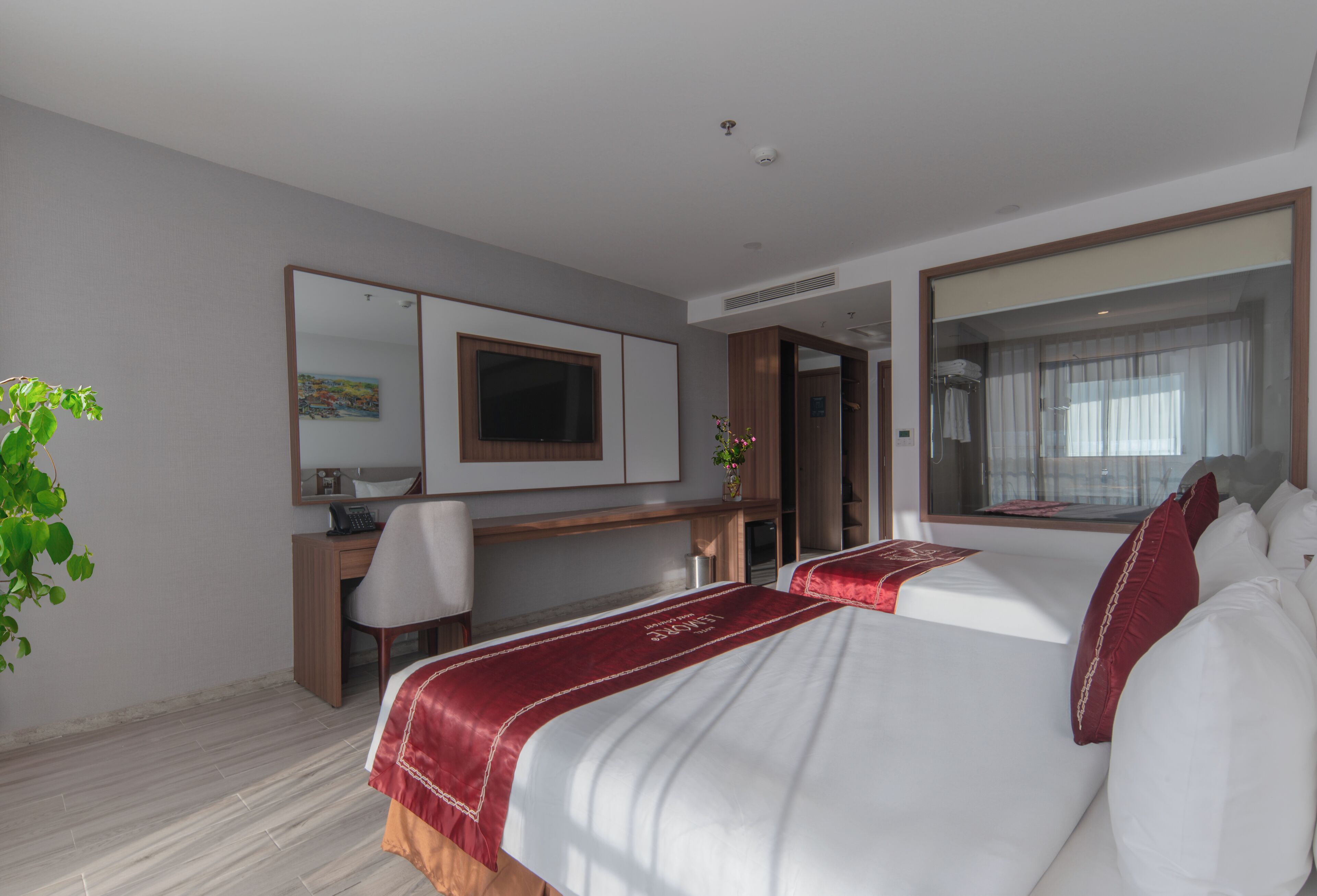 A hotel room with two beds, a desk, a chair, a mirror, and a plant.