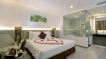 A hotel room with a neatly made bed, a sitting area with a chair and a small table, a large mirror, and a bathroom with a glass shower enclosure.