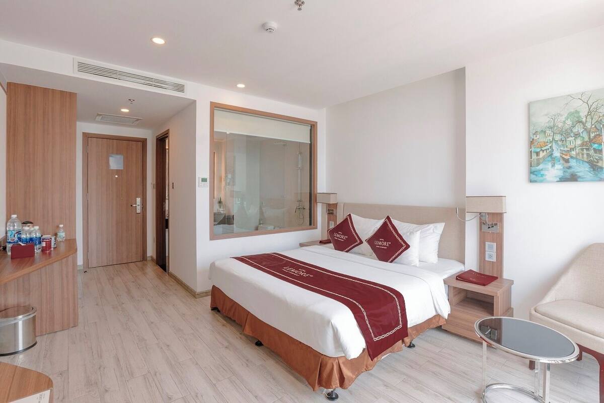 A hotel room with a large bed, a bedside table, a chair, a painting on the wall, and a bathroom visible through an open door.