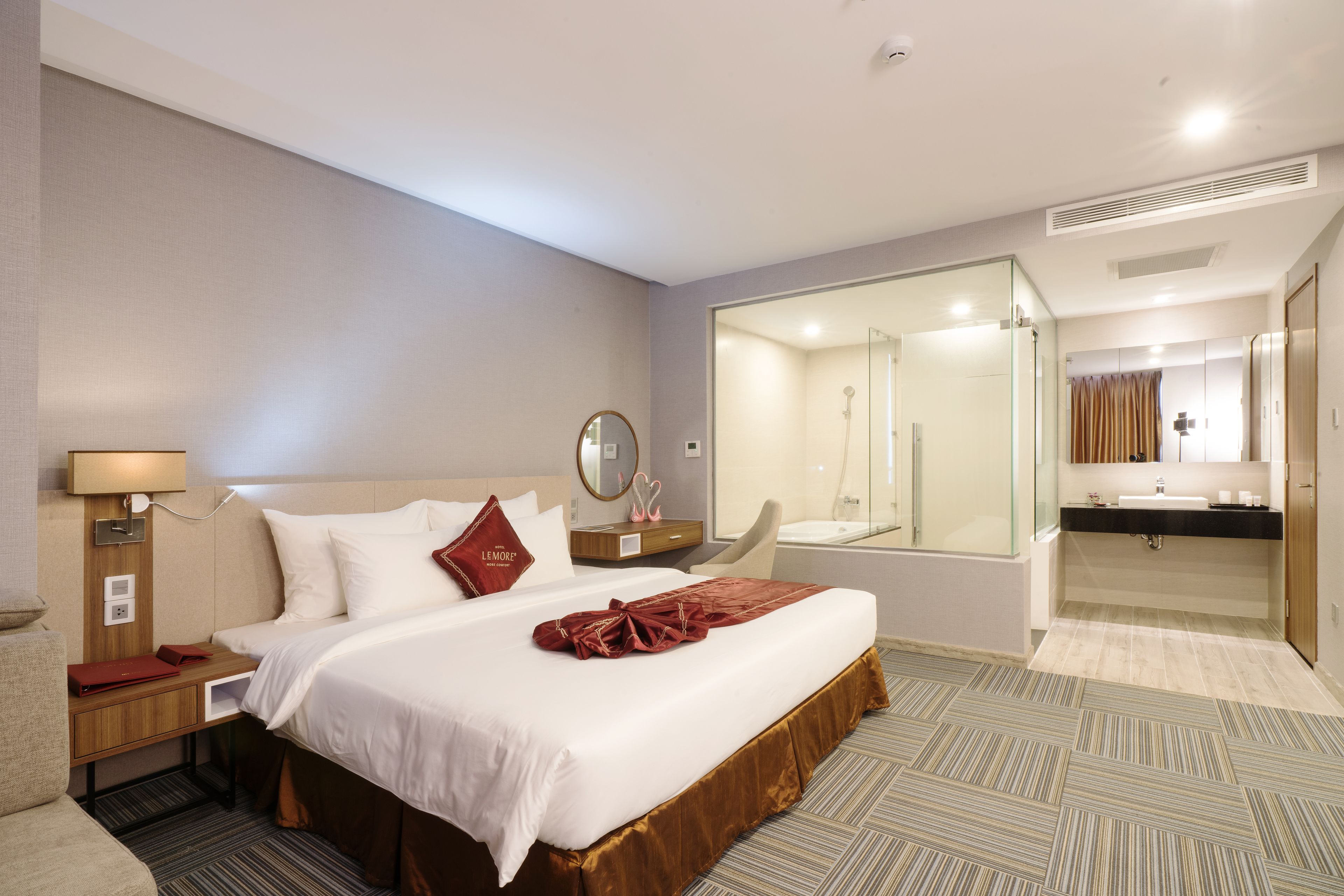 A hotel room with a large bed, a bedside table with a lamp, a mirror, a bathroom with a sink and mirror, and a shower area.