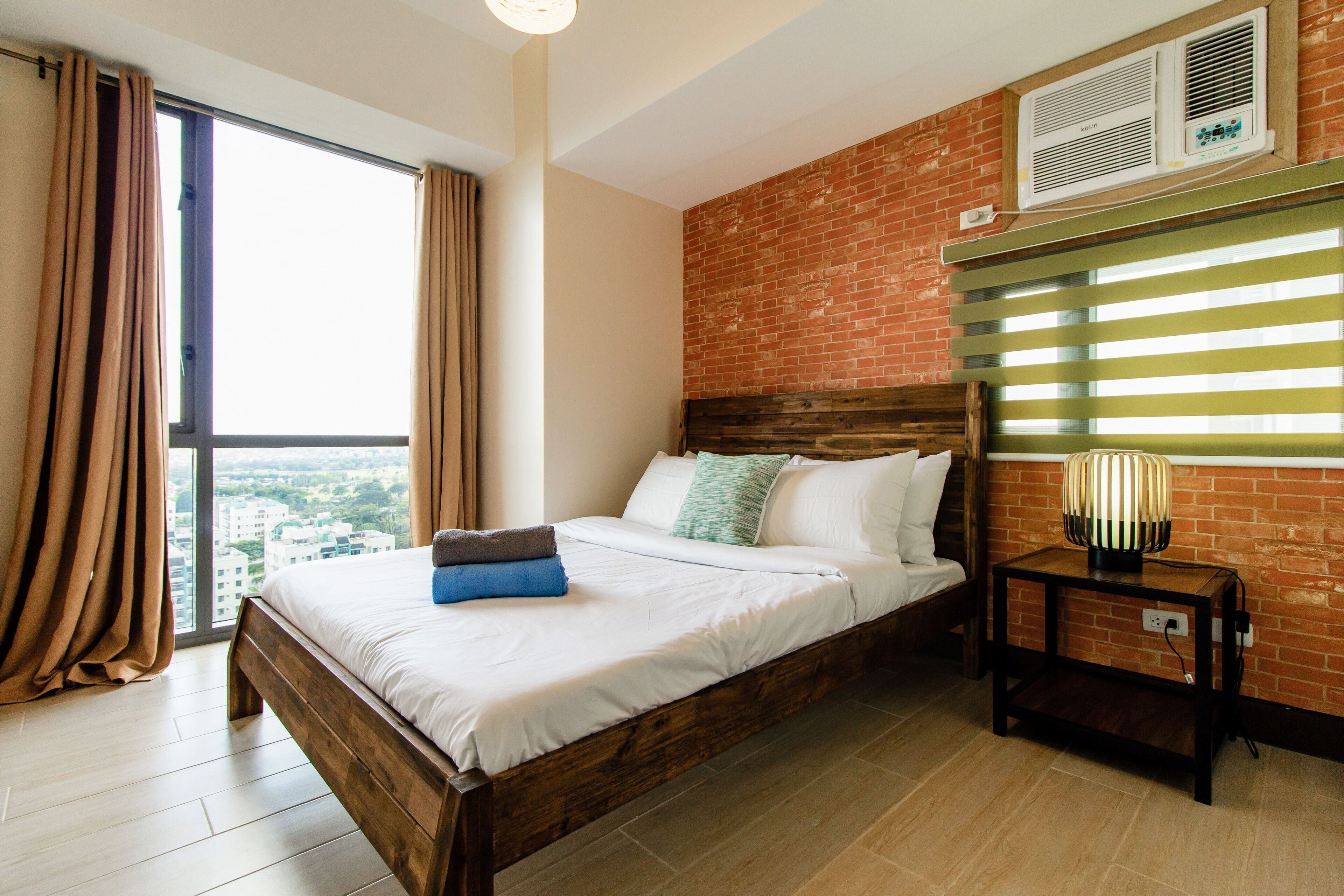 A bedroom with a large window, a brick wall, a wooden bed, and a nightstand with a lamp.