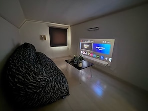 A living room with a sofa, a small table with plants, a wall-mounted TV displaying various apps, and a window with blinds.