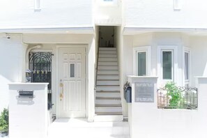 A white building with a front door, a staircase leading to the entrance, and a small garden area with a metal fence. - Soshigaya Apartment (Tokyo)