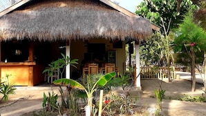 A thatched-roof building with a covered outdoor seating area, surrounded by tropical vegetation. - Ings Garden (Gili Air)