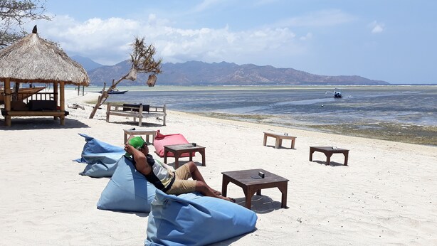 A beach with a thatched-roof hut, lounge chairs, and a wooden table. - Ings Garden (Gili Air)