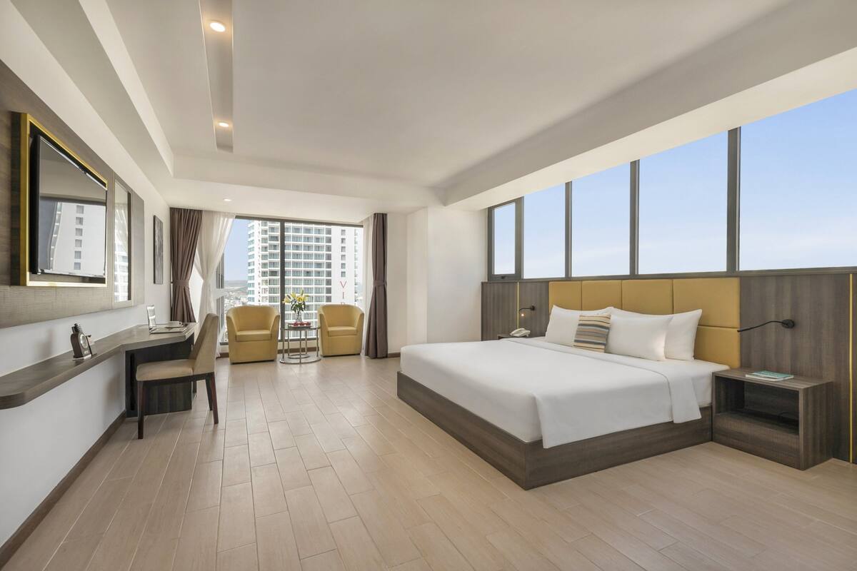 Une chambre d’hôtel moderne dotée d’un grand lit, d’un bureau avec une chaise, d’une petite table avec un vase et d’une grande fenêtre donnant sur la ville.