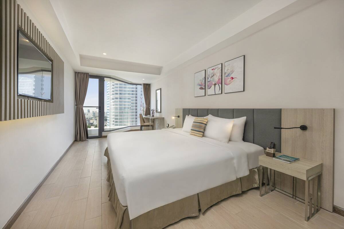 Une chambre d’hôtel moderne dotée d’un grand lit, d’une table de chevet, d’un bureau et offrant une vue sur la ville grâce à de grandes fenêtres.