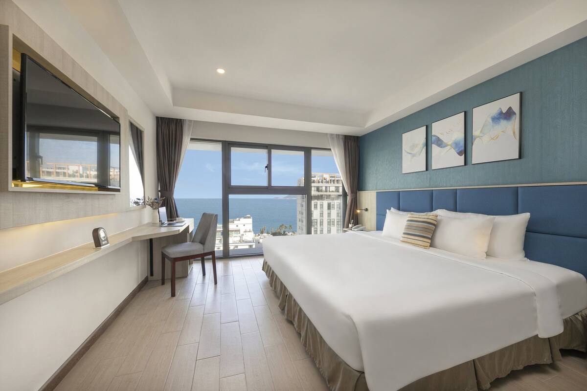 Une chambre d’hôtel avec un grand lit, un bureau avec une chaise, un téléviseur fixé au mur et une vue sur la mer.