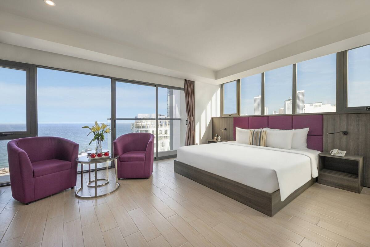 Une chambre d’hôtel moderne avec un grand lit, deux fauteuils, une petite table agrémentée de fleurs et une vue sur la ville grâce à de grandes fenêtres.