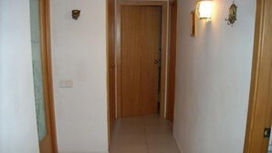 Un pasillo estrecho con una puerta de madera al final, paredes blancas y un accesorio de iluminación en la pared derecha.