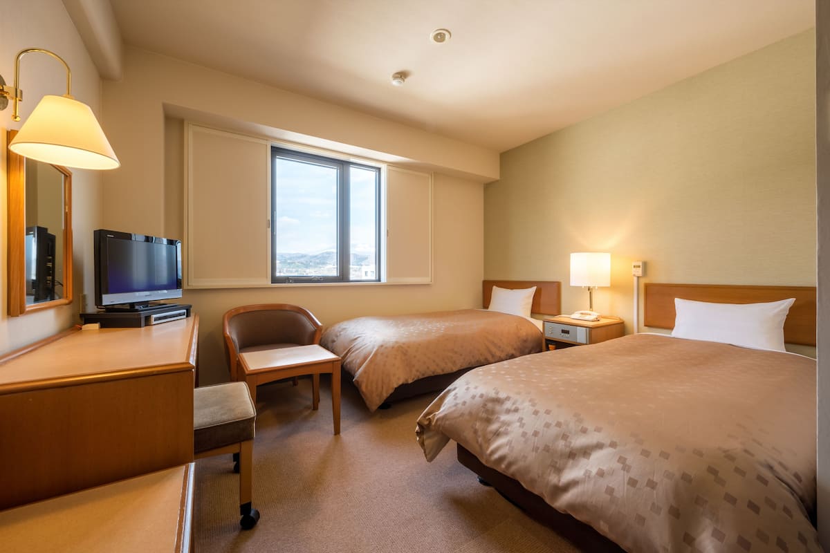 A hotel room with two beds, a desk with a television, a chair, a window with a view, and a lamp.