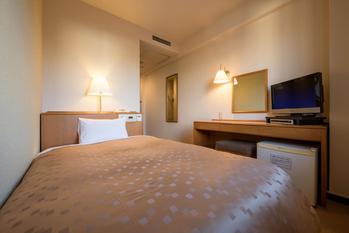 A hotel room with a bed, a desk with a television, a lamp, and a small refrigerator.
