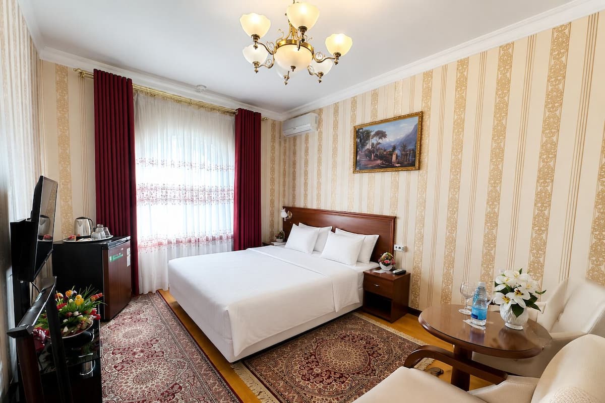 A hotel room with a large bed, a desk with a computer, a chair, a small table with a vase of flowers, and a painting on the wall.