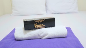 Een keurig opgevouwen witte handdoek op een paarse handdoek, met een zwarte doos met de tekst 'Kamar Keluargo' op een bed.