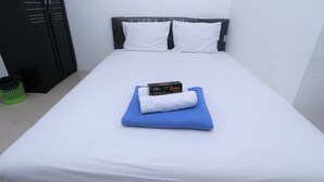 Een net opgemaakt bed met witte lakens, een blauw kussen en een gevouwen handdoek. Op het bed staat een zwart doosje, mogelijk een boek of een afstandsbediening. Links van het bed staat een groene prullenbak.