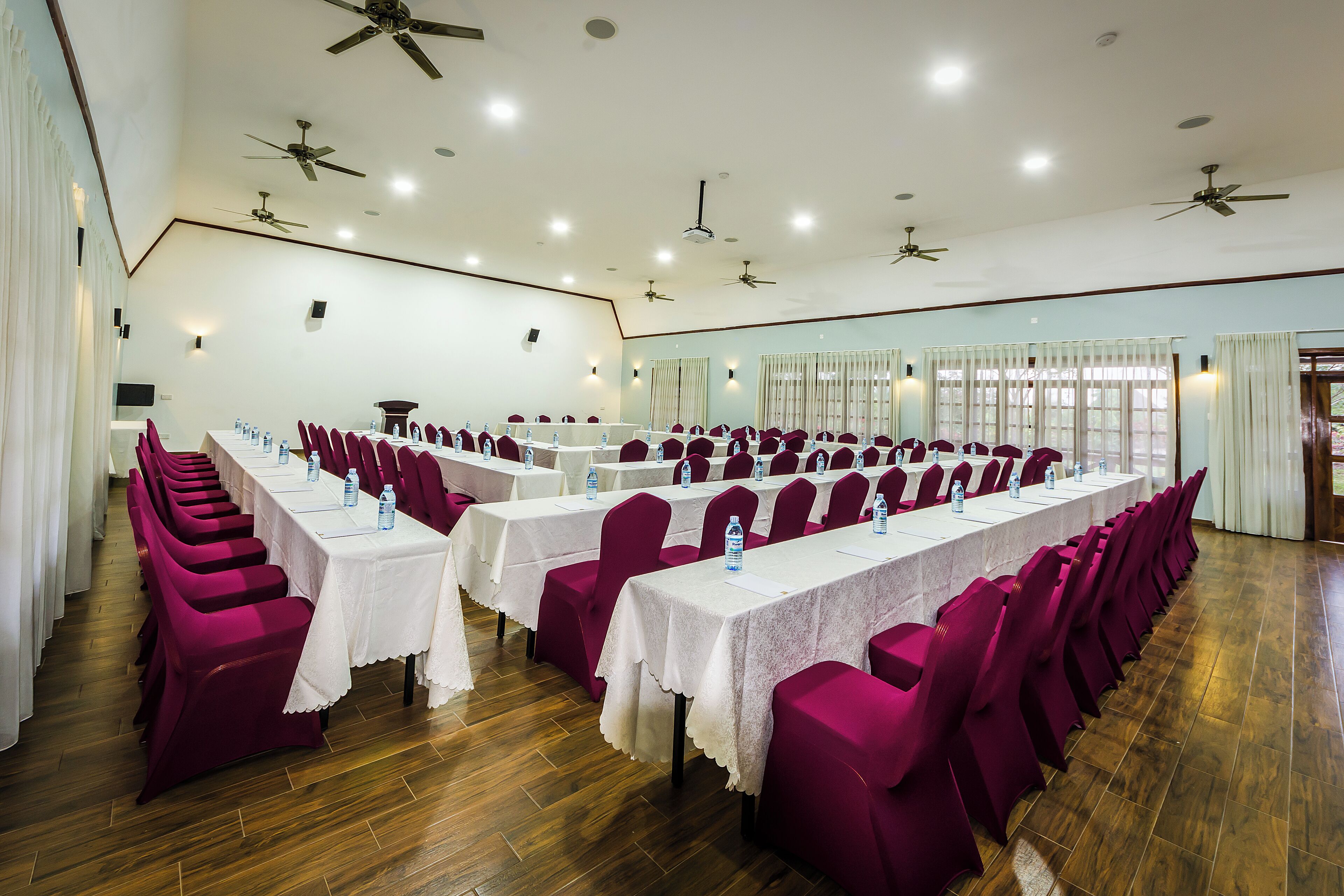A conference room with rows of tables and chairs, ceiling fans, and windows with curtains.
