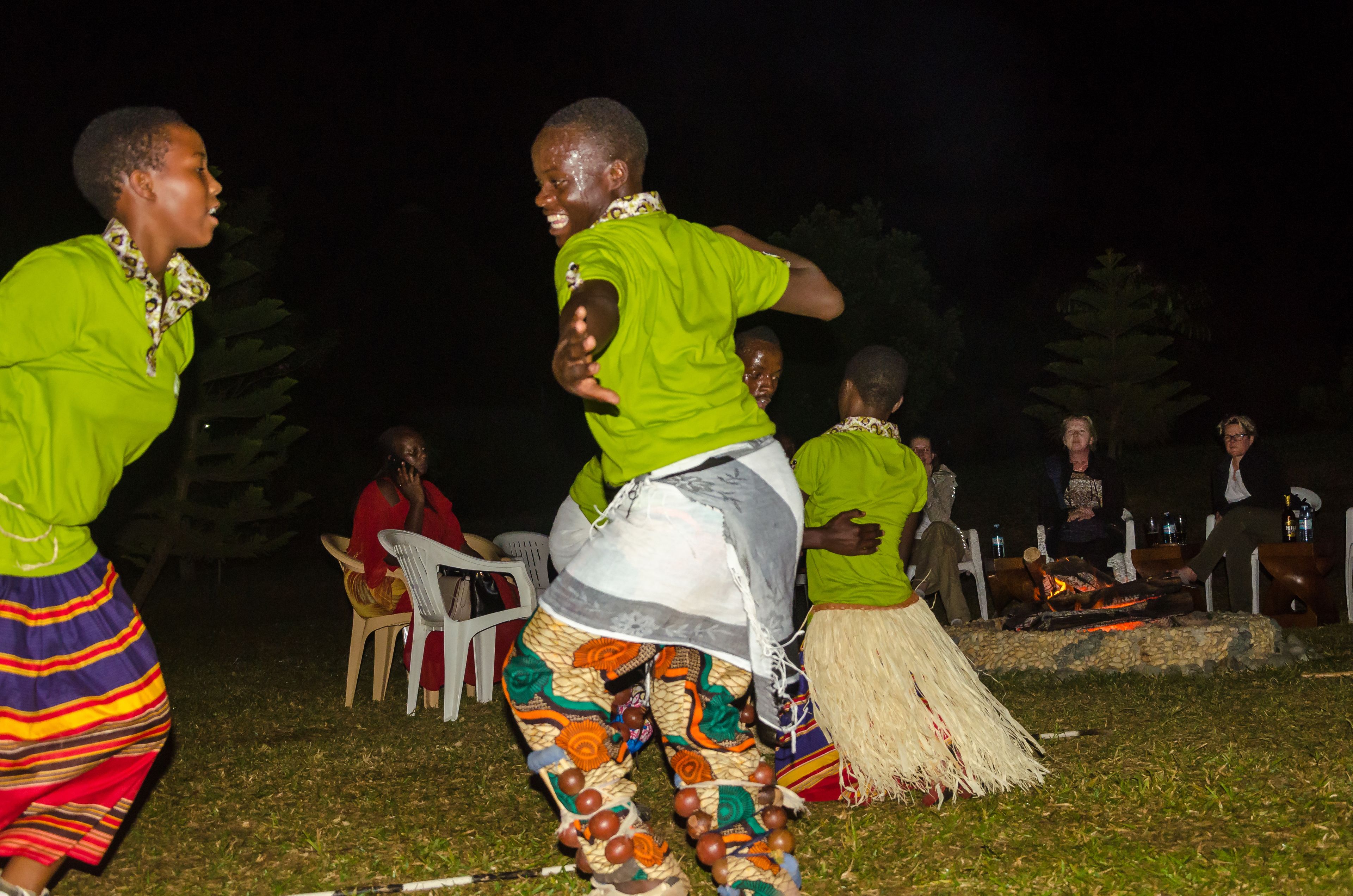 A group of people outdoors at night, some wearing traditional attire, with a fire and plastic chairs in the background.