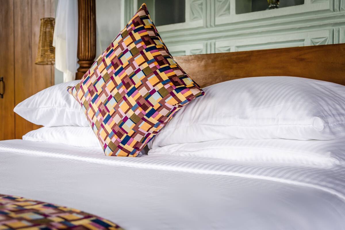 A bed with white linens and a colorful woven pillow.