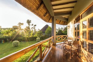 Una terraza de madera con techo de paja, barandillas de madera y vistas a un jardín exuberante y una montaña al fondo.