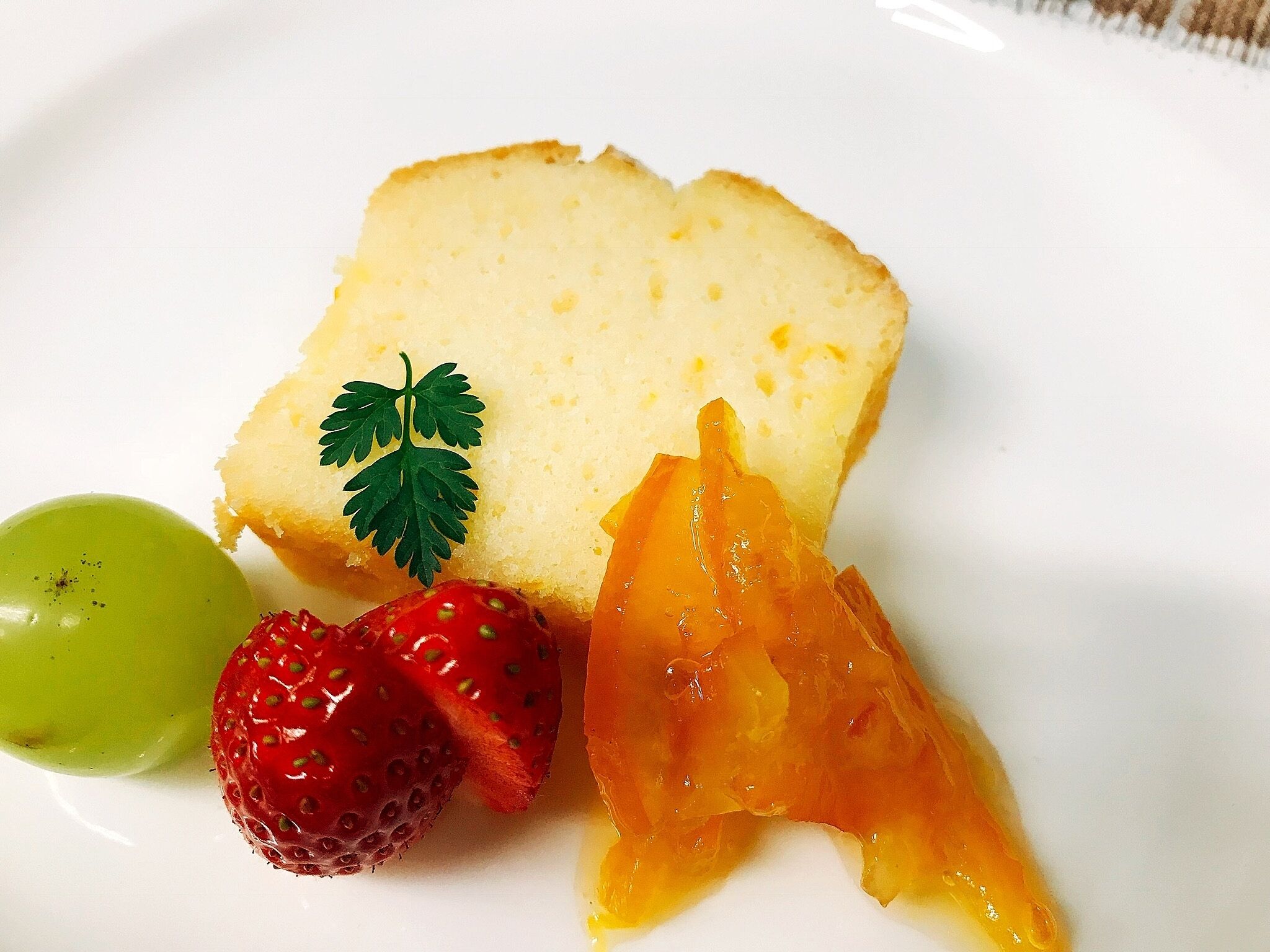 A slice of cake with a sprig of parsley, a green grape, and a strawberry on a white plate.