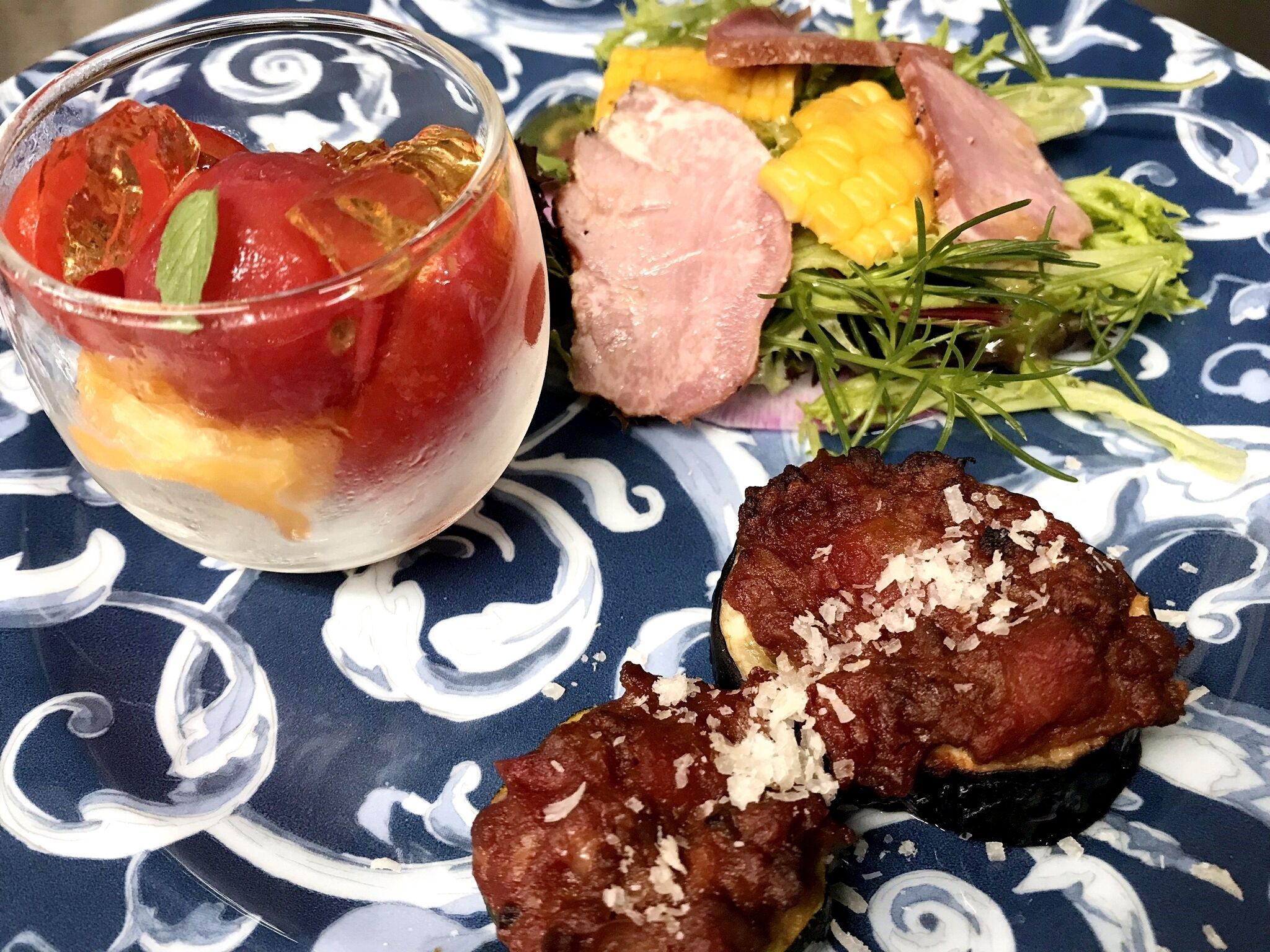 Une assiette contenant une portion de soupe à la tomate, garnie d’herbes, une salade composée de légumes verts et de maïs, et un morceau de viande recouvert de fromage râpé.
