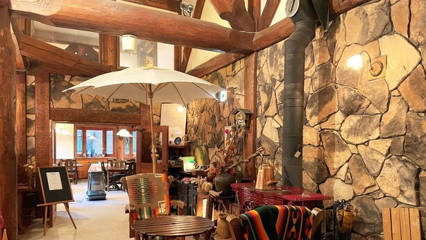 Interior rústico com paredes de pedra, vigas de madeira e lareira de pedra. Há cadeiras de madeira e uma mesa com um cobertor colorido.