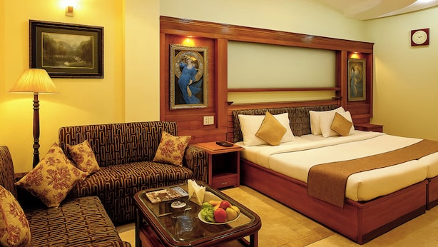 Une chambre d’hôtel avec un grand lit, un canapé, une table basse avec des fruits et des œuvres d’art encadrées accrochées aux murs.