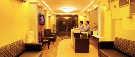 A hotel reception area with a reception desk, two sofas, a chandelier, and a staircase.