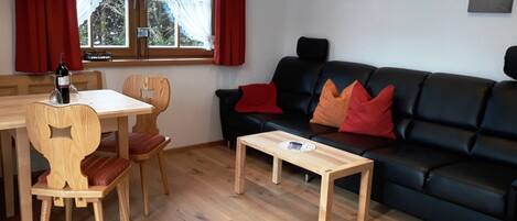 A living room with a black sofa, wooden chairs, a wooden table, and a window with curtains.