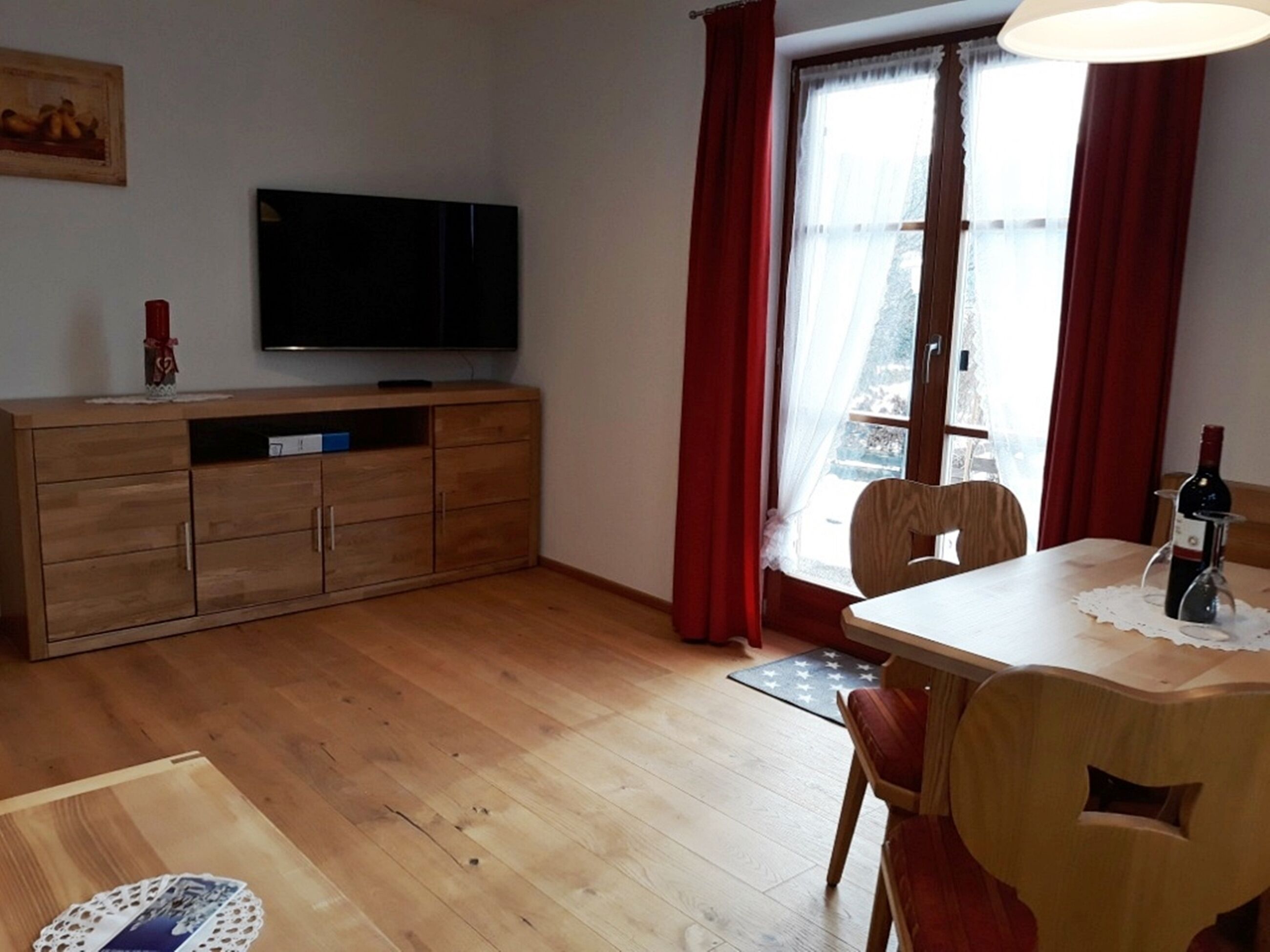 Un salon avec un meuble en bois, un téléviseur, une table à manger avec des chaises et une fenêtre ornée de rideaux rouges.