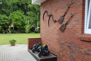 A brick building with a metal sculpture of gardening tools on the wall and a paved area with a potted plant and parked motorcycles.