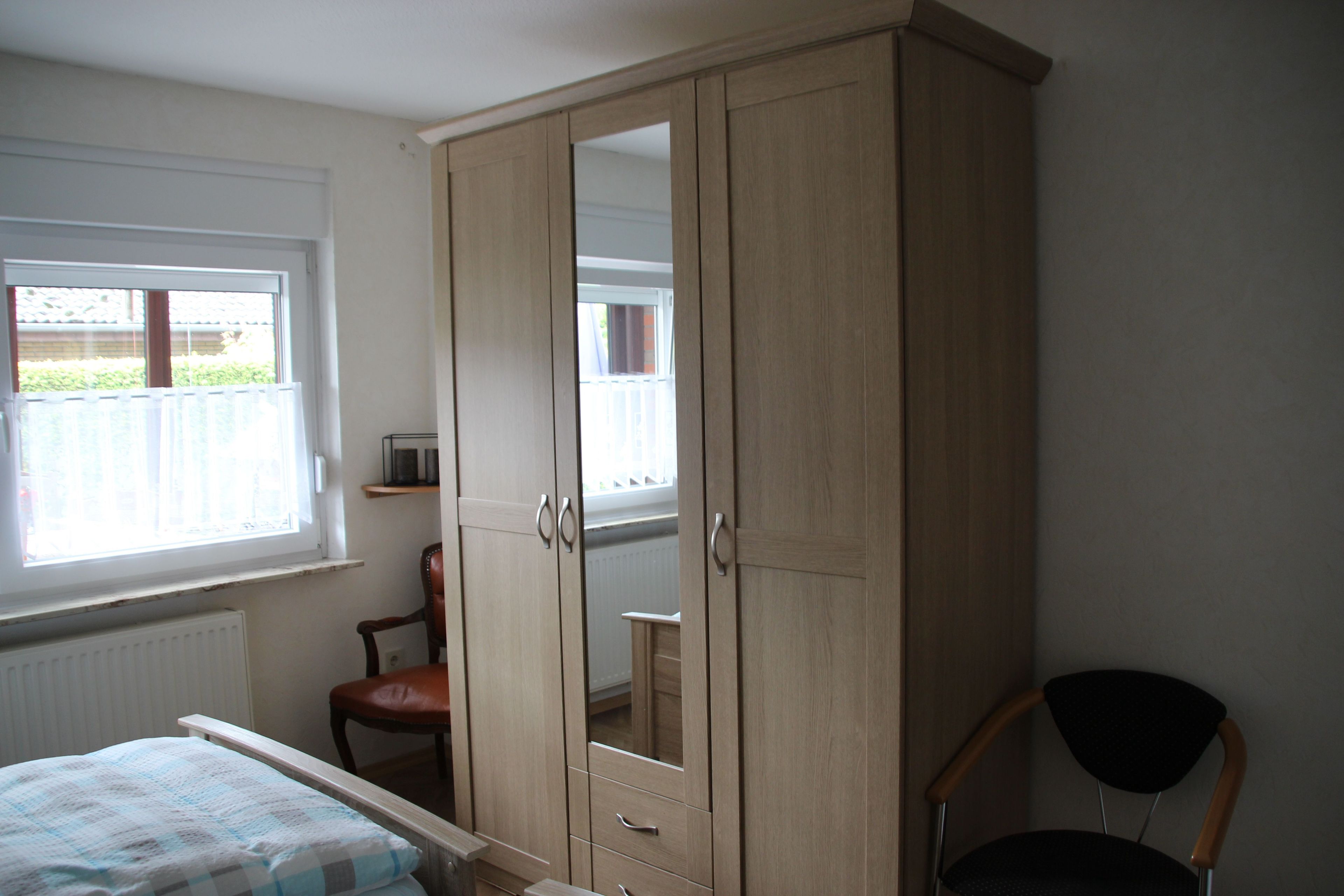 Een slaapkamer met een grote kast, een bed met een geruit beddegoed, een stoel en een raam met uitzicht op een hek.
