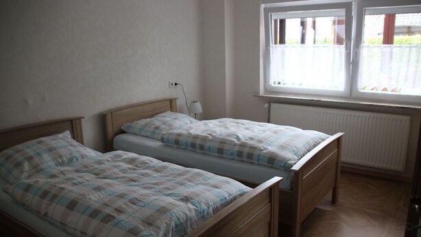 Un dormitorio con dos camas de madera, una ventana con cortinas blancas y un radiador.