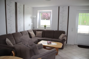 A living room with a sectional sofa, a wooden coffee table, and a window with a view of greenery. - FeWo - Apartment Drieling, 85091 (Ostrhauderfehn)