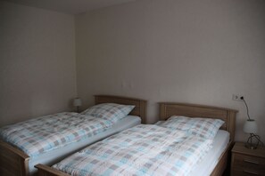 A room with two single beds, each with a wooden headboard and a checkered bedspread. There is a bedside table with a lamp on the right side.