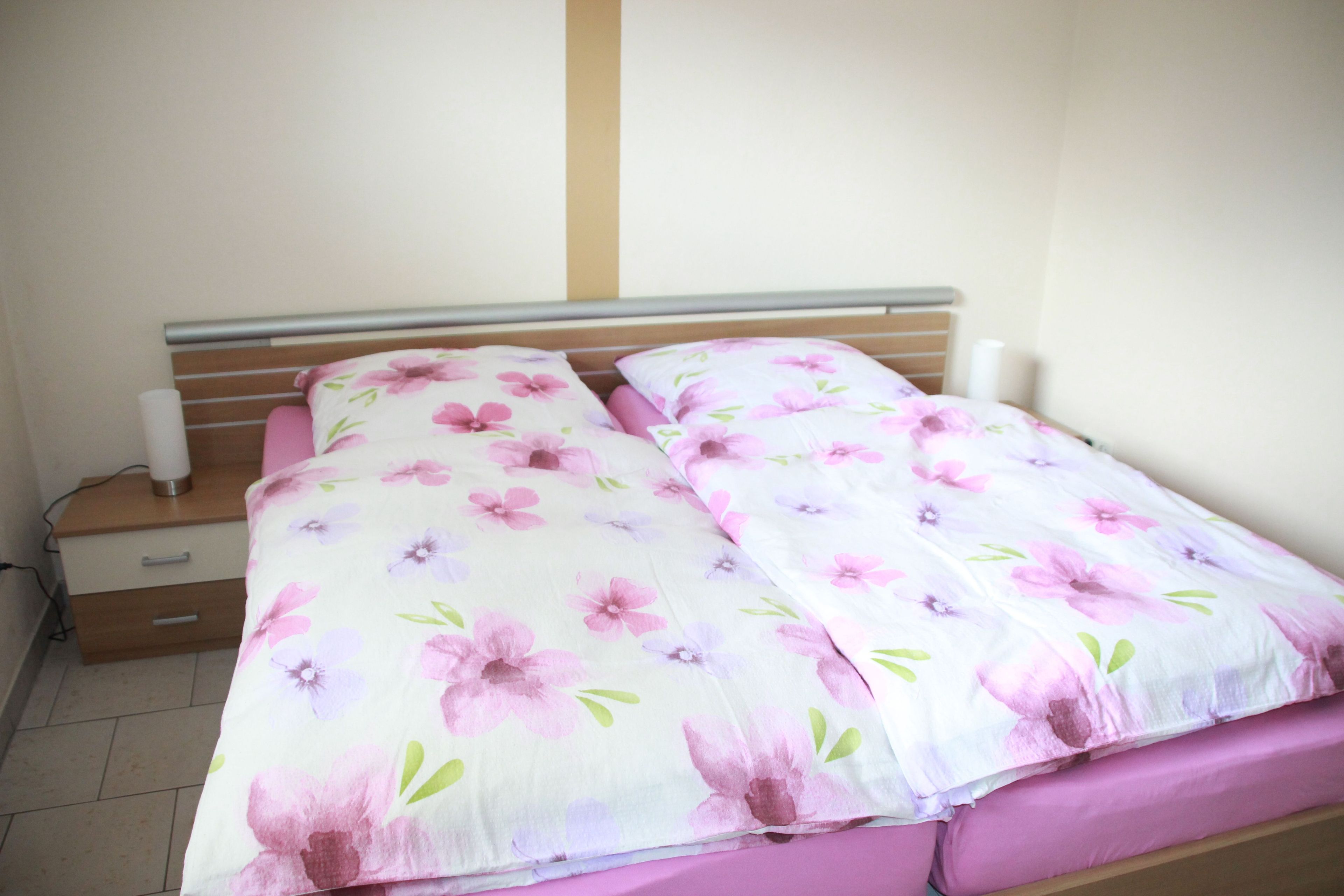 Una cama con una colcha estampada con flores y dos almohadas, una mesita de noche de madera con una lámpara y un tablero con cajones.