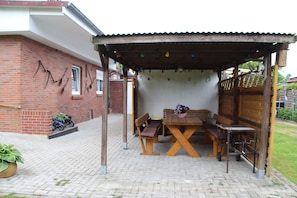 A covered outdoor seating area with a wooden table and benches, a small table with a flower pot, and a brick building in the background. - FeWo - Apartment Drieling, 85091 (Ostrhauderfehn)