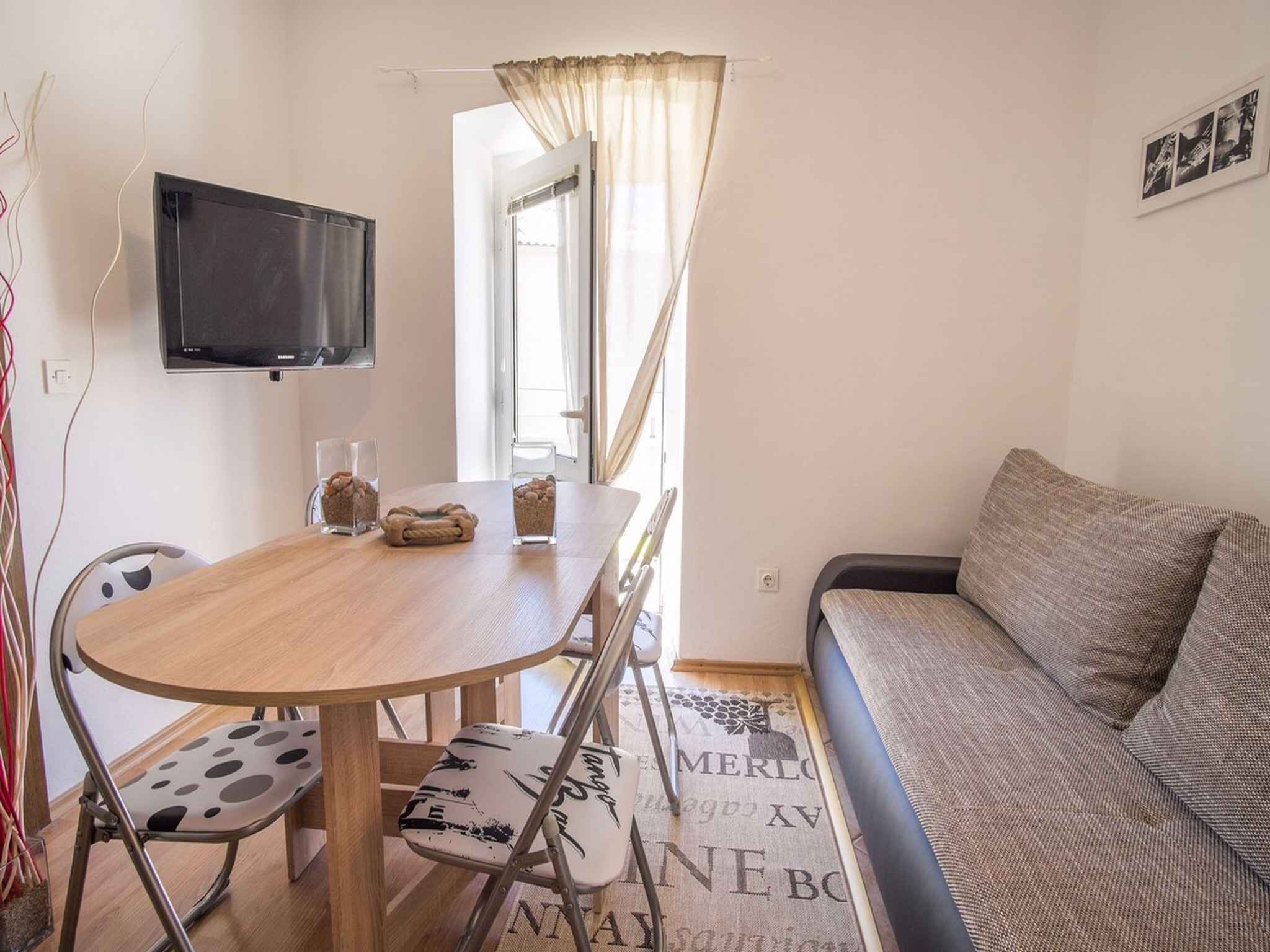 Un espace de vie compact comprenant une table à manger en bois, deux chaises, un canapé, un téléviseur à écran plat et une fenêtre avec des rideaux.