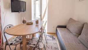 Een knus woongedeelte met een houten eettafel, twee stoelen, een bank, een flatscreen televisie en een raam met gordijnen.
