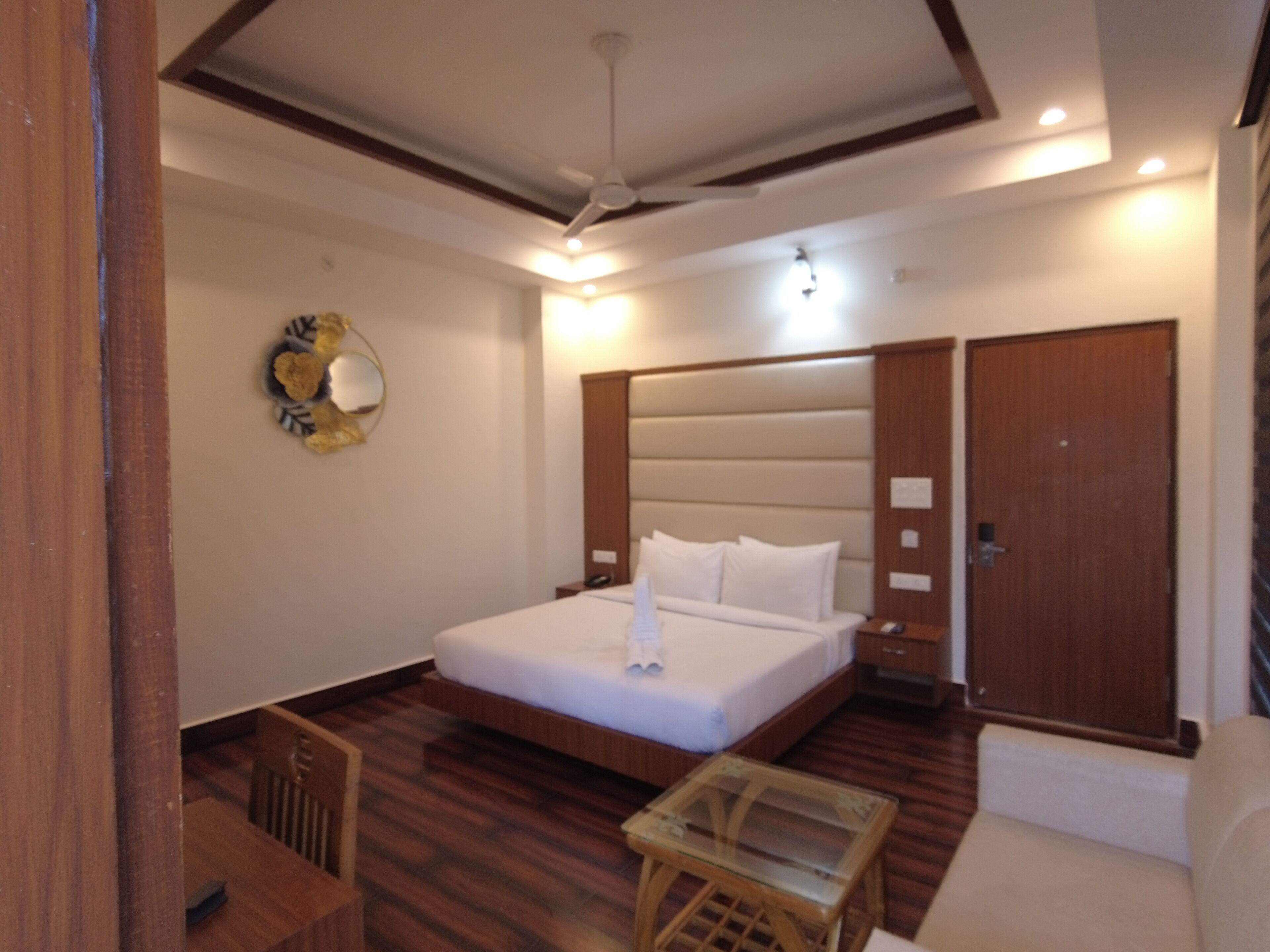 A modern hotel room with a large bed, a wooden headboard, a glass table, and a wooden floor.