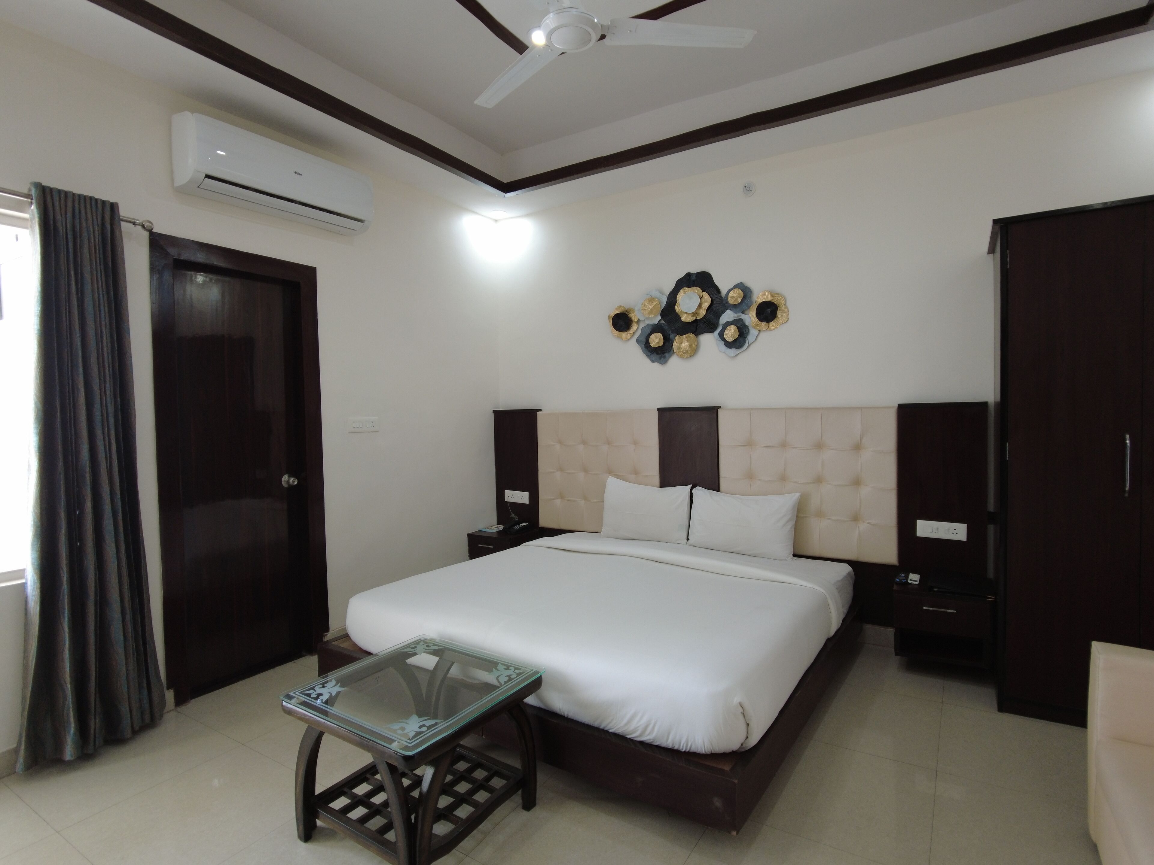 Une chambre d’hôtel comprenant un lit, une table en verre, un ventilateur de plafond, un appareil de climatisation et un élément décoratif mural.