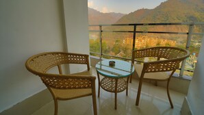 A balcony with wicker chairs and a glass table, overlooking a mountainous landscape. - Kamay The Grand Alova (Narendranagar)