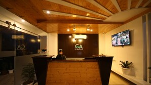 A reception desk with a wooden finish and a logo, a flat-screen TV on the wall, and potted plants. - Kamay The Grand Alova (Narendranagar)